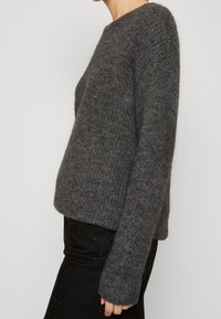 Grau gestrickter Pullover mit rundem Ausschnitt und langen Ärmeln, der einen strukturierten Stoff und eine lockere Passform zeigt. Kombiniert mit einem schwarzen, strukturierten Rock.