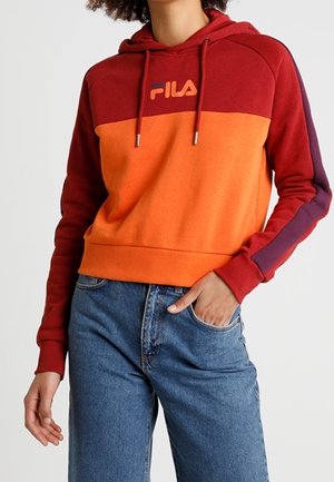 Woman wearing a red and orange FILA hoodie with drawstrings and blue jeans, one hand in pocket, against a plain background.