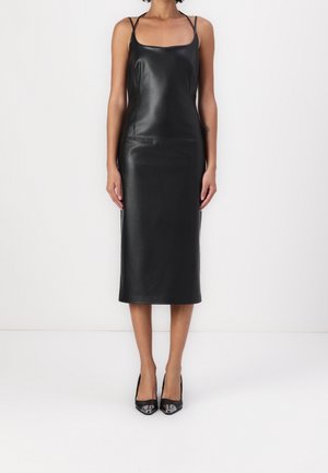 Frau, die ein ärmelloses schwarzes Leder-Midikleid mit dünnen Trägern und schwarzen spitz zulaufenden High Heels trägt, steht vor weißem Hintergrund.