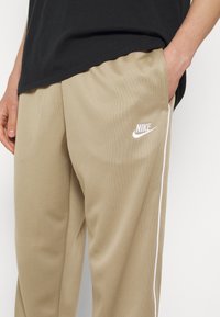 Pantalon de sport beige clair en tissu texturé, avec un logo Nike blanc et des bandes latérales pour plus de détails.