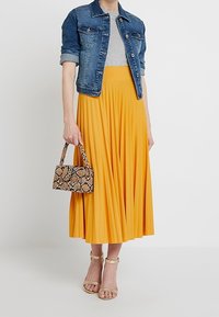 Chaqueta de mezclilla, camiseta gris, falda amarilla plisada y fluida, y sandalias de tacón beige; bolso pequeño de estampado de serpiente con asa.