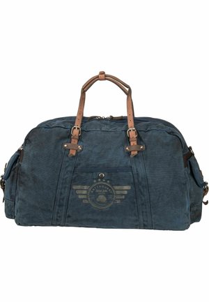 Blue canvas duffel bag with brown leather accents, featuring a front logo design, two handles, and zipped side pockets.