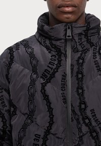 Black puffer jacket with a high collar, featuring a quilted design. It has a glossy finish and prominent chain and text patterns. Zip closure.