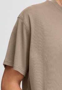 Camiseta corta de algodón color tostado con un corte relajado, que presenta una superficie texturizada y costuras visibles a lo largo de las mangas.