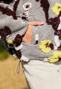 Gray fleece sweatshirt with floral applique in maroon, white, and yellow. Features a front pocket and adjustable drawstring at the hem.