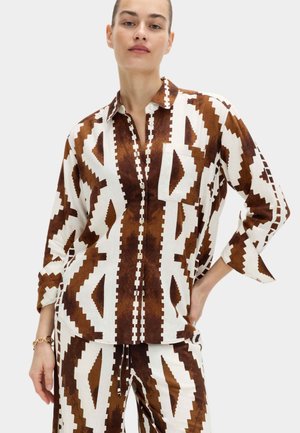 Jeune femme portant une chemise boutonnée à motifs géométriques blancs et bruns et un pantalon assorti à coupe décontractée, main sur la hanche.