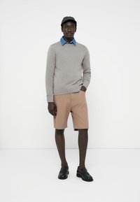 Grauem Pullover, hellblauem Hemd mit Kragen, beigen Shorts mit strukturiertem Muster, schwarzen Slippern und einer schwarzen Mütze. Schlichter Hintergrund.