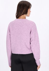 Lila kortärmad tröja med en mjuk, fluffig textur. Har sänkt axel och långa ärmar samt en ribbad nederkant. Bärs tillsammans med svarta byxor.
