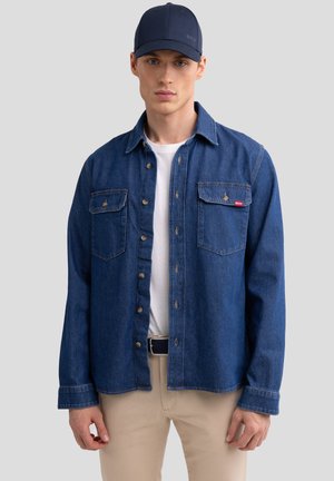 Chemise en denim bleu foncé avec fermeture boutonnée, deux poches poitrine et coutures contrastantes ; assortie à un t-shirt blanc et un pantalon beige.