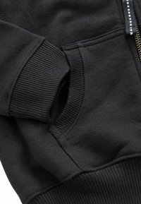 Schwarze Reißverschluss-Hoodie mit gerippten Bündchen, einer Fronttasche und einer glatten Textur. Detail in der Nähe des Reißverschlussbereichs sichtbar.