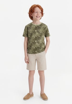 Roodharige jongen die lacht, draagt een groen t-shirt met bladpatroon, beige shorts en lichtbruine instapschoenen, en staat met één hand in zijn zak.