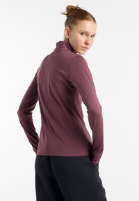 Langarm T-shirt in donker mauve, gemaakt van een glad, rekbaar materiaal. Kenmerken zijn een hoge kraag en een aansluitend ontwerp, gecombineerd met zwarte broeken.