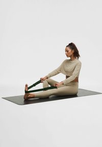 Jasno beżowy krótki top i dopasowane legginsy, siedząc na ciemnej macie do jogi, używając zielonego pasa do rozciągania, aby sięgnąć jedną stopą.