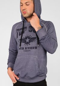 Lila huvtröja gjord av mjukt material, med en framficka och ett grafiskt tryck med vit och svart text samt stjärnsymbol.