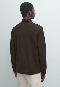 Brązowa koszula z długim rękawem i kołnierzykiem. Tkanina wydaje się fakturowana, o prostym kroju i mankietach na guziki. Tło jest jednolite, jasnoszare.