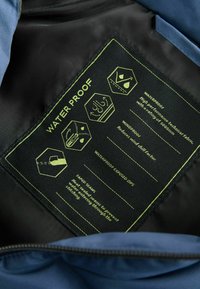 Étiquette à l'intérieur de la veste bleue détaillant le tissu imperméable, les caractéristiques coupe-vent, les coutures scellées, et les zips étanches avec des icônes pour chacun.
