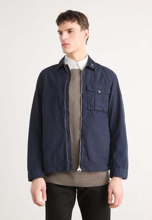 Veste zippée bleu marine avec un col haut, une poche à l'avant et une doublure intérieure. Portée par-dessus un pull beige et un pantalon noir, présentant une texture lisse.