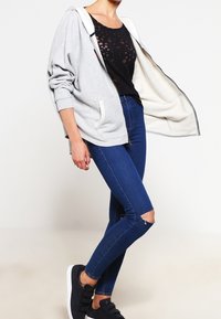 Šedá mikina na zip s bílým vnitřkem, kombinovaná s černým topem s květinovým vzorem a modrými skinny džíny s dírami na kolenou. Černé tenisky.