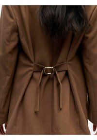 Bruine jas met een gladde textuur, voorzien van een tailledetail met een zilveren gesp en vastgemaakte stoffen banden aan de achterkant.