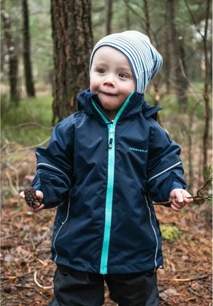 Blue waterproof jacket with teal zipper and accents, paired with black pants. Child holds a pine cone and a small branch, wearing a striped hat.