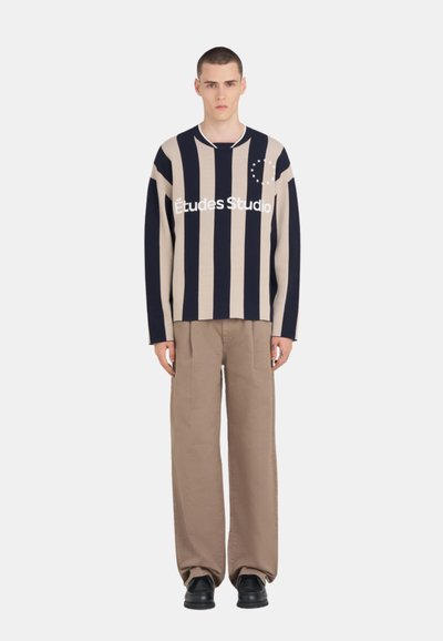 Homme debout portant un pull à rayures verticales bleu marine et beige avec le texte "Études Studio" et un pantalon ample beige avec des chaussures noires.