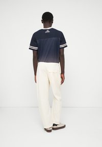 T-shirt de sport bleu marine avec des rayures blanches sur les manches et un pantalon large couleur crème. Présente un logo au dos et une poche sur le pantalon.