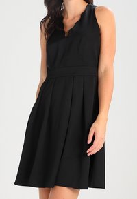 Black sleeveless dress with a v-neckline, fitted bodice, and pleated skirt. Made from smooth fabric with no visible patterns or accents.