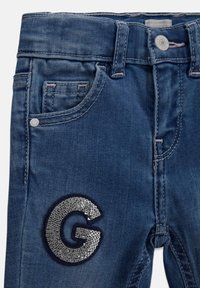 Denim jeans in medium blue with a smooth texture. Features a decorative silver sequin "G" patch and traditional five-pocket design.