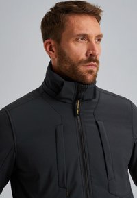 Chaqueta gris oscuro con un cuello alto, con cierre de cremallera, múltiples detalles en panel y una textura mate. Marcada con "PME Legend."