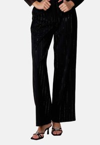 Pantalons noirs à jambes larges avec des rayures verticales brillantes. Tissu doux avec une texture lisse. Associés à des sandales noires ornées de bijoux.