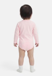 Tout-petit aux cheveux foncés, debout de dos, portant un body à manches longues rose clair et des chaussettes blanches, sur un fond blanc.