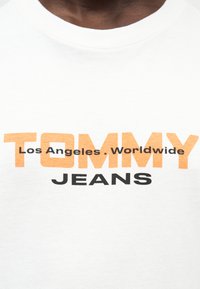 Λευκό βαμβακερό t-shirt με έντονο μαύρο και πορτοκαλί κείμενο: "TOMMY" στο πάνω μέρος, "Los Angeles. Worldwide" πάνω από το "JEANS" στο κάτω μέρος.