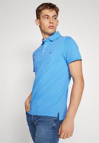 Polo bleu en coton, doté d'un col, d'une patte de boutonnage à trois boutons et de fentes sur les côtés. Broderie du logo sur la poitrine.