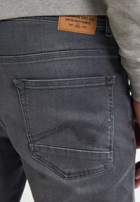 Vaqueros de denim gris oscuro con un acabado texturizado. Presenta un parche de cuero en la cinturilla y un detalle de costura curvada en el bolsillo trasero.