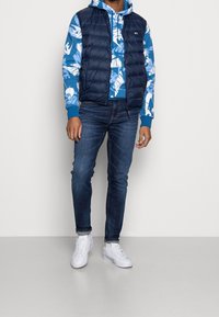 Námořnická péřová vesta přes modrou mikinu s bílým listovým vzorem, kombinovaná s tmavými džíny a bílými teniskami. Vyznačuje se různorodými texturami a barvami.