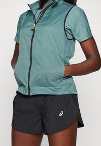 Leichte, türkisfarbene Zip-Jacke mit perforierten Details und schwarzen Akzenten, kombiniert mit schwarzen Shorts mit weißem Logo.