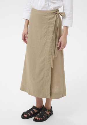 Mujer con una falda beige envuelta atada a la cintura, combinada con sandalias negras de tejido y una camisa blanca de manga larga, de pie sobre un fondo blanco.