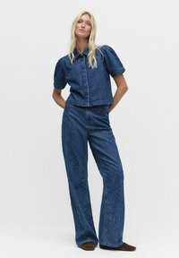 Vrouw staat met handen achter haar rug en draagt een blauwe spijkerblouse met puffmouwen en bijpassende wijde jeans, met bruine schoenen.