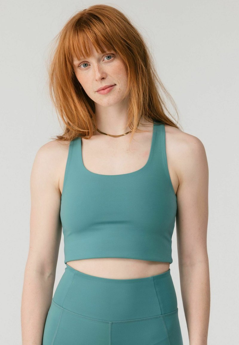 Vörös hajú nő zöldeskék haspólóban és magas derekú leggingsben, egyszínű világos háttér előtt áll, előre nézve.