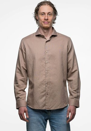 Man met lichtbruin haar en korte baard, die een beige overhemd met knopen en blauwe spijkerbroek draagt, staand tegen een effen witte achtergrond.