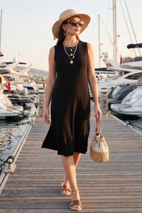 Mujer con vestido negro sin mangas, sombrero de paja, gafas de sol y sandalias caminando por un muelle de una marina sosteniendo un bolso marrón claro y beige al atardecer.