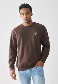 Man in een bruine sweatshirt met wolflogo en de tekst "BRAVE SPIRIT", gecombineerd met lichtblauwe jeans, staand tegen een effen achtergrond.