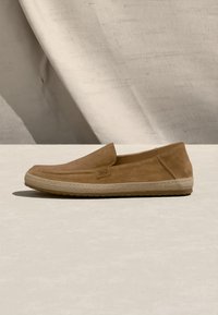 Φωτεινό καφέ σουέτ παπούτσι τύπου slip-on με πλεκτή μπεζ σόλα, επίπεδο τακούνι και μινιμαλιστικό σχέδιο, τοποθετημένο σε ουδέτερο υφασμάτινο φόντο.