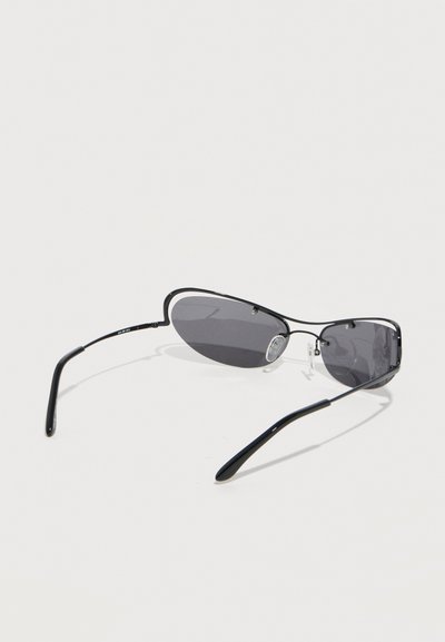 Μαύρα στενά οβάλ γυαλιά ηλίου με λεπτό μεταλλικό πλαίσιο και σκούρους φακούς, εμφανιζόμενα να ακουμπούν σε λευκή επιφάνεια.