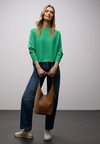 Femme portant un pull vert, un jean bleu et des baskets blanches, tenant un sac à main en suède marron devant un fond gris uni.