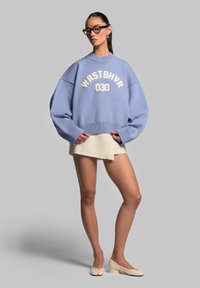 Maglione oversize di colore azzurro chiaro lavorato a maglia con lettere bianche "WRSTBHV" e "030" sulla parte anteriore. Abbinato a una gonna beige e scarpe color crema.