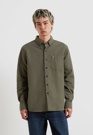 Giovane con capelli corti e con riflessi chiari, indossa una camicia verde oliva con bottoni e jeans blu scuro, in piedi davanti a uno sfondo semplice.