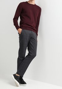 Burgunderfarbener Strickpullover kombiniert mit grauen Karohosen. Das Outfit wird durch schwarze Sneakers mit weißen Sohlen ergänzt.