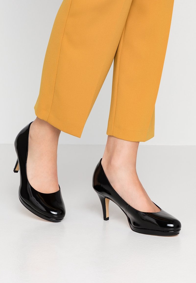 Jambes portant un pantalon jaune moutarde et des escarpins noirs brillants à talons moyens, debout sur un sol blanc lisse.