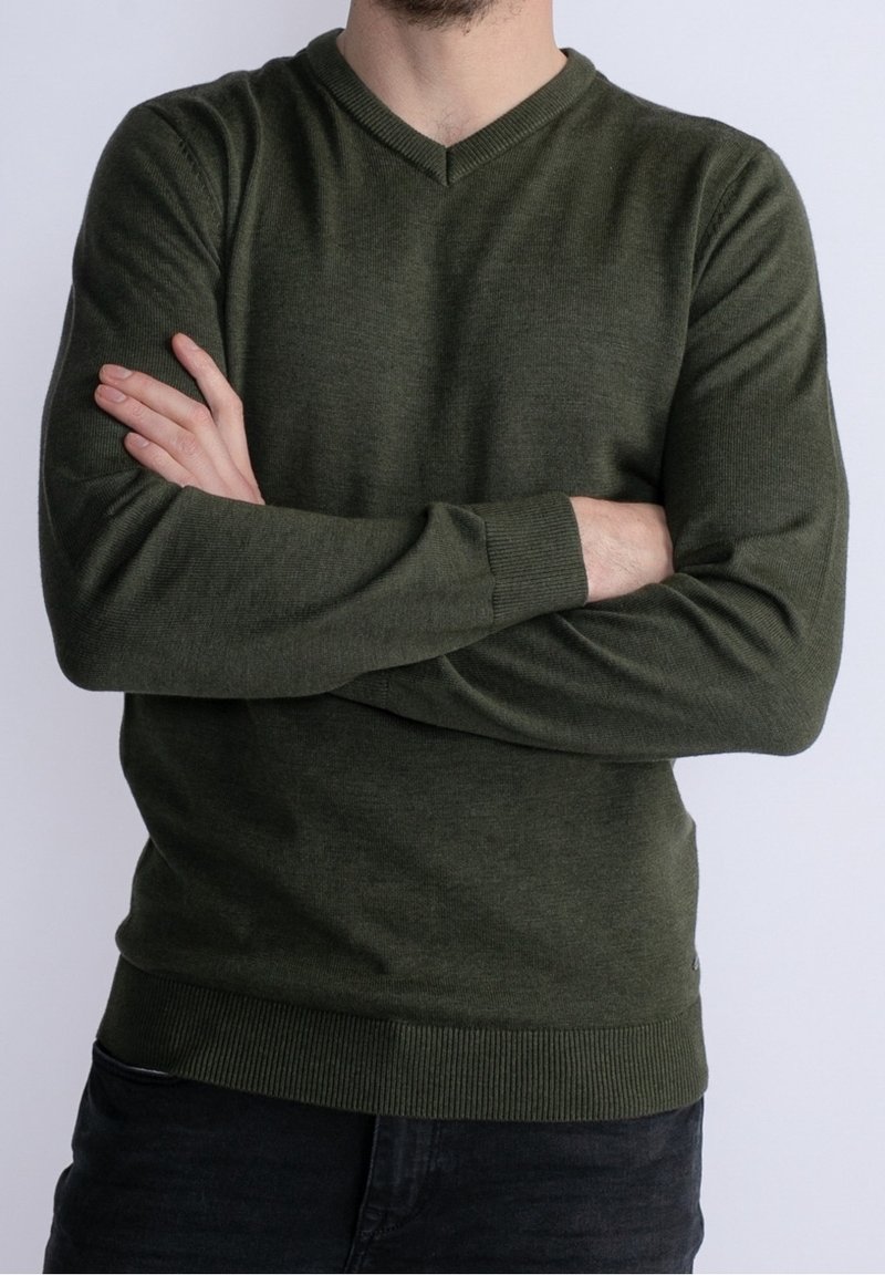 Homme portant un pull à col en V vert foncé, les bras croisés, sur un fond clair uni.